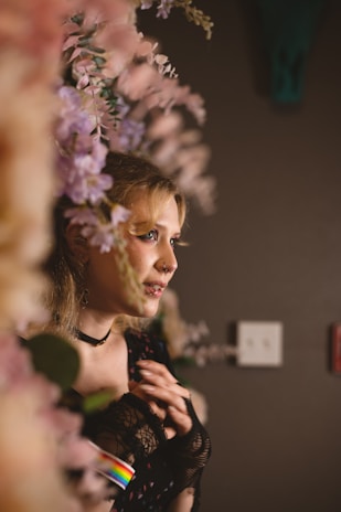 A person with blonde hair and facial piercings stands amidst a backdrop of pink and purple flowers. The lighting casts warm tones, and the person wears a lacy black outfit with a choker. A subtle rainbow detail is visible on their sleeve.