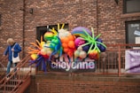 A colorful balloon arrangement with various shapes and sizes is displayed against a brick wall. A person in a blue jacket and jeans is walking down a nearby staircase holding onto the railing. There are windows with signage and a banner displaying text behind the balloons.