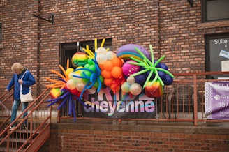 A colorful balloon arrangement with various shapes and sizes is displayed against a brick wall. A person in a blue jacket and jeans is walking down a nearby staircase holding onto the railing. There are windows with signage and a banner displaying text behind the balloons.