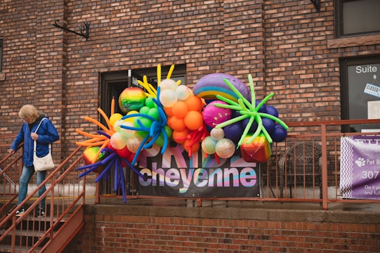 A colorful balloon arrangement with various shapes and sizes is displayed against a brick wall. A person in a blue jacket and jeans is walking down a nearby staircase holding onto the railing. There are windows with signage and a banner displaying text behind the balloons.