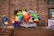 A colorful balloon arrangement with various shapes and sizes is displayed against a brick wall. A person in a blue jacket and jeans is walking down a nearby staircase holding onto the railing. There are windows with signage and a banner displaying text behind the balloons.