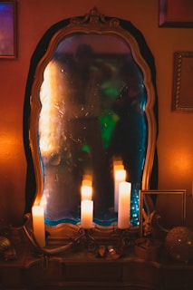 An ornate mirror reflecting a dimly lit room with warm and cool lighting. Several candles are placed on a surface in front of the mirror, their flames casting a soft glow. Decorative items surround the mirror, including a small figurine and a spherical ornament. The atmosphere suggests a cozy and mystical setting.