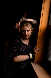 A person with light hair stands in a dimly lit space, leaning against a wooden surface by a window. They wear a dark, patterned dress and fishnet gloves, with their makeup accentuating their eyes. A wristband with a rainbow pattern is visible. The lighting creates high contrast, emphasizing their pose and expression.