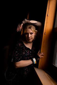A person with light hair stands in a dimly lit space, leaning against a wooden surface by a window. They wear a dark, patterned dress and fishnet gloves, with their makeup accentuating their eyes. A wristband with a rainbow pattern is visible. The lighting creates high contrast, emphasizing their pose and expression.