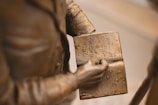 A close-up view of a bronze statue depicting a person holding an open book. The engraved text on the book includes phrases such as 'CHARTING THE TERRITORY 1867' and 'OPENING THE AMERICAN WEST.' The statue's detailed craftsmanship highlights the folds of the figure's clothing.