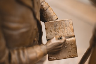 A close-up view of a bronze statue depicting a person holding an open book. The engraved text on the book includes phrases such as 'CHARTING THE TERRITORY 1867' and 'OPENING THE AMERICAN WEST.' The statue's detailed craftsmanship highlights the folds of the figure's clothing.