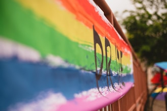 A brightly colored banner featuring a horizontal rainbow pattern with the word 'PRIDE' partially visible. The vibrant colors are set against a blurry background, suggesting an outdoor setting with trees. The image focuses on the texture and bold colors of the banner.