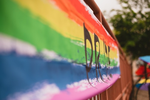 A brightly colored banner featuring a horizontal rainbow pattern with the word 'PRIDE' partially visible. The vibrant colors are set against a blurry background, suggesting an outdoor setting with trees. The image focuses on the texture and bold colors of the banner.