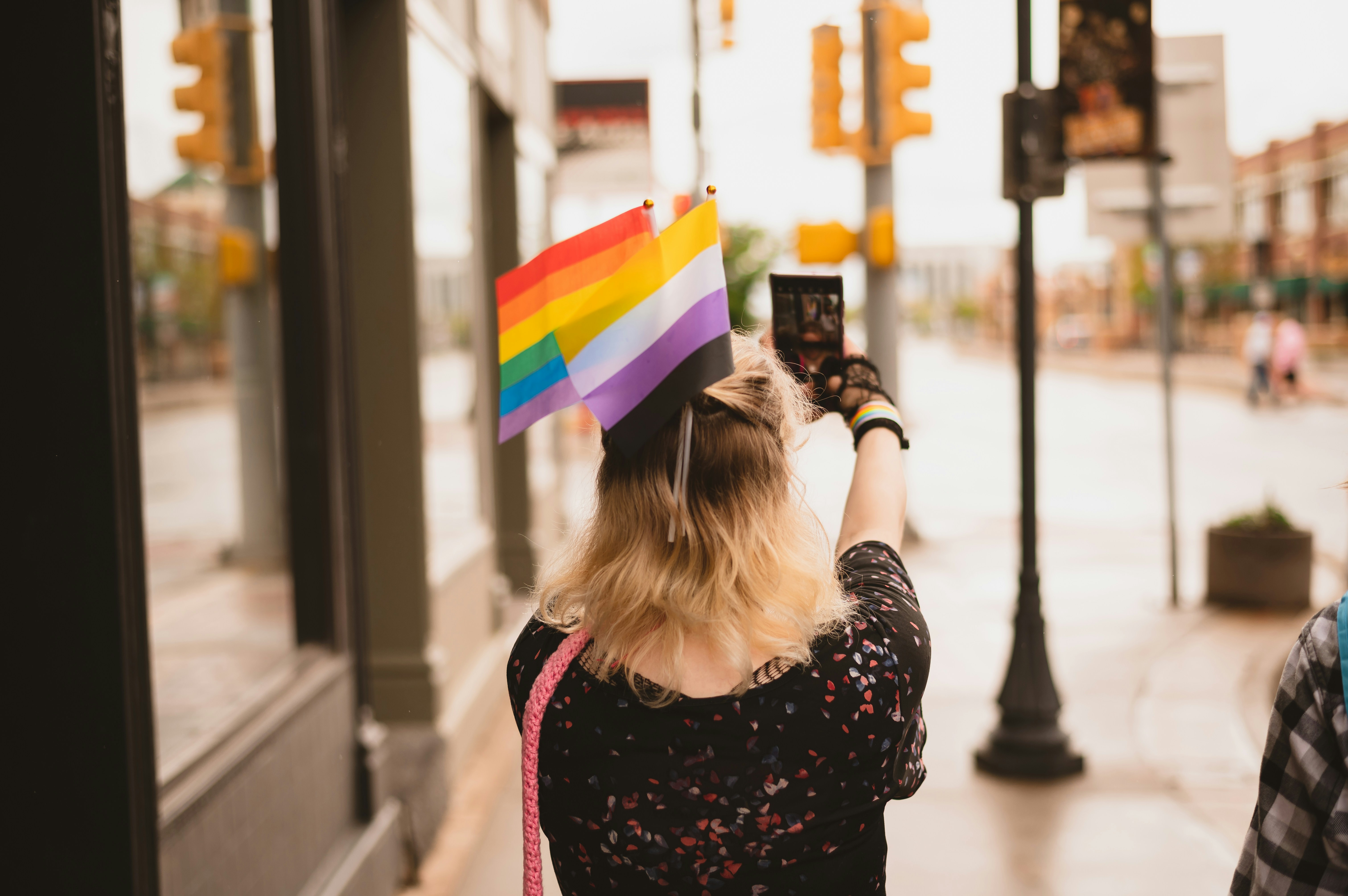 A stylish person with two Pride flags in their hair holds up a smartphone to take a picture on a city street.
