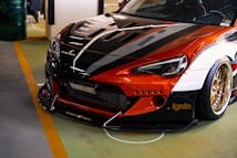A customized sports car with a sleek, aerodynamic design featuring a mix of black, orange, and white colors. The front view highlights the aggressive styling with a prominent grille, detailed headlights, and a low-profile bumper. The car is equipped with gold alloy wheels and detailed decals and branding on the hood and body.