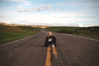 A person is sitting on the middle of a long, empty road that stretches into the distance. The landscape is open with rolling hills on either side under a partly cloudy sky. The road has a yellow dividing line and no traffic, symbolizing solitude and freedom.