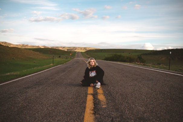 A person is sitting on the middle of a long, empty road that stretches into the distance. The landscape is open with rolling hills on either side under a partly cloudy sky. The road has a yellow dividing line and no traffic, symbolizing solitude and freedom.