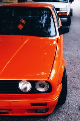 An orange vintage car with prominent headlights and a sleek design is parked on a street. The focus is on the front portion of the car, with an orange pair of dice hanging inside the windshield. A white car is parked behind the orange car, slightly blurred.