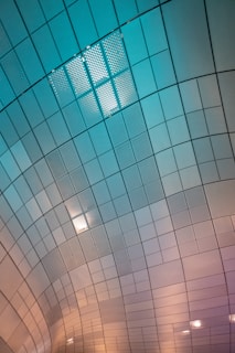 A geometrically patterned ceiling consisting of several rectangular tiles arranged in a curved, wave-like structure. The tiles exhibit a gradient of colors from teal to pink, giving a dynamic and modern appearance. Some tiles have perforated designs, and there are integrated light fixtures providing illumination.