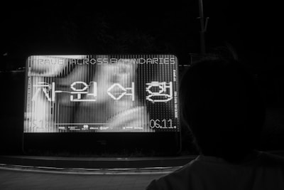 A person is standing in front of a large illuminated billboard at night. The billboard displays text which appears to be in a foreign language, with digital-like distortions across it. The surrounding environment is mostly dark, emphasizing the brightness of the billboard.
