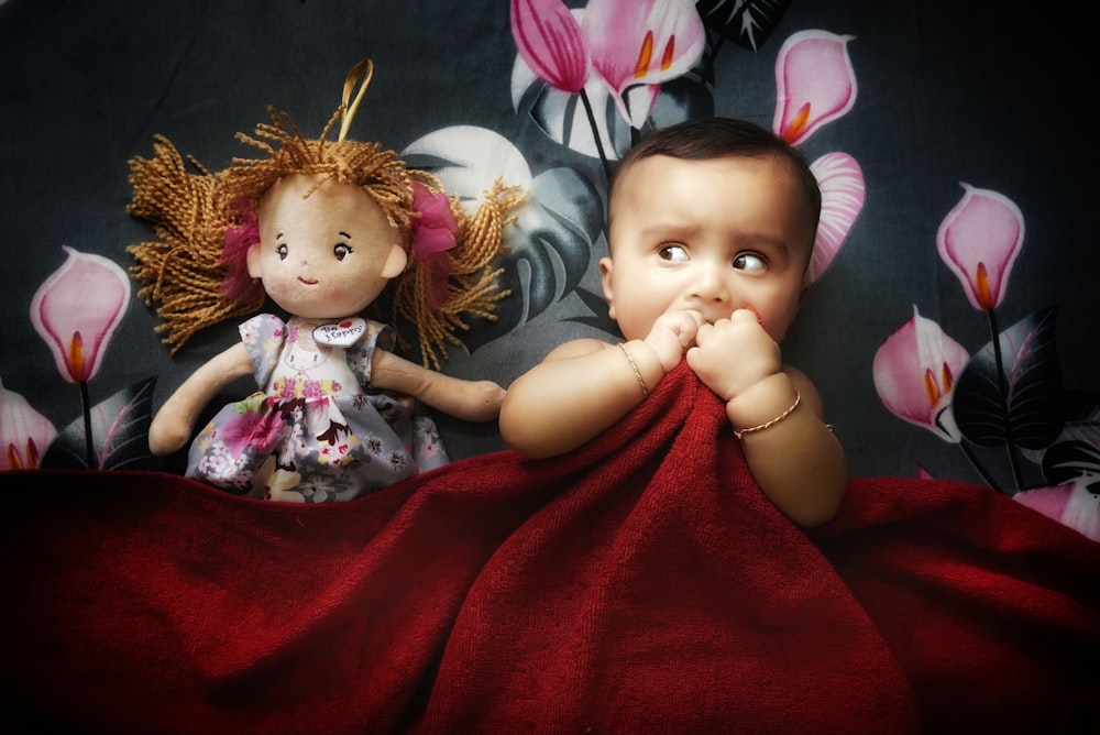 a baby and a doll laying on a red blanket