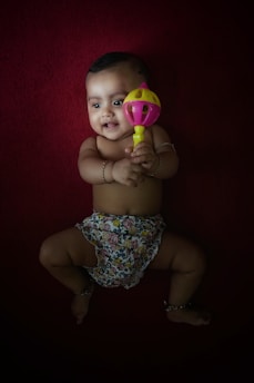 A baby lying on a deep red background, holding a brightly colored rattle. The baby is wearing floral-patterned shorts and has a joyful expression. A couple of bracelets adorn the baby's wrists and ankles.