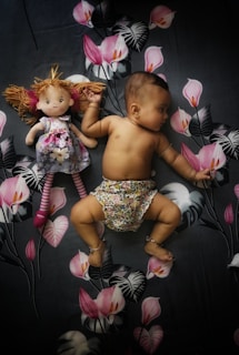 A baby lies on a floral-patterned fabric, wearing patterned shorts. Next to the baby is a doll with a floral dress and striped leggings. The background features pink flowers and large leaves, creating a playful and cozy setting.