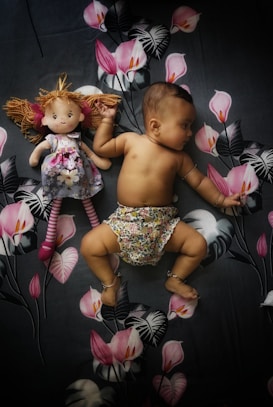 A baby lies on a floral-patterned fabric, wearing patterned shorts. Next to the baby is a doll with a floral dress and striped leggings. The background features pink flowers and large leaves, creating a playful and cozy setting.
