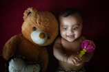 A baby lying on a dark red surface, smiling and holding a colorful rattle. Next to the baby is a large brown teddy bear, adding a cozy and playful element.