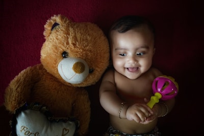 A baby lying on a dark red surface, smiling and holding a colorful rattle. Next to the baby is a large brown teddy bear, adding a cozy and playful element.
