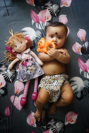 A baby lying on a floral patterned blanket, holding an orange toy in their hand. Beside the baby is a doll with curly hair, wearing a floral dress and striped leggings. The baby is dressed in floral-patterned shorts and adorned with bracelets on their wrist and ankle.