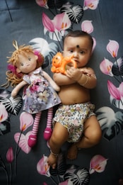 A baby lying on a floral patterned blanket, holding an orange toy in their hand. Beside the baby is a doll with curly hair, wearing a floral dress and striped leggings. The baby is dressed in floral-patterned shorts and adorned with bracelets on their wrist and ankle.