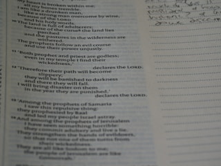 Hands writing on a notebook with a Bhagavad Gita open nearby.