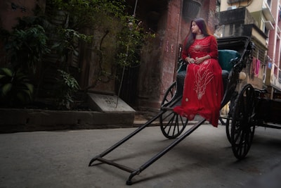 A woman in a red dress sits on a traditional hand-pulled rickshaw in a narrow alleyway. The surrounding environment features old buildings with overgrown plants and weathered walls, creating an atmosphere of nostalgia. The woman appears contemplative, adding a serene mood to the scene.