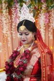A person dressed in traditional wedding attire adorned with intricate gold jewelry and a red and gold saree. The headpiece is ornate and white, adding to the elegance. The backdrop features floral decorations and soft lighting, enhancing the ceremonial atmosphere.