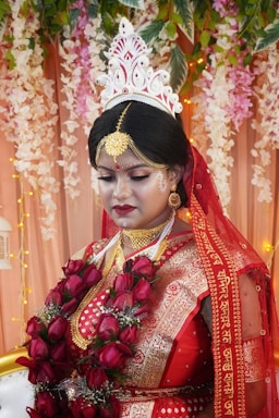 A person dressed in traditional wedding attire adorned with intricate gold jewelry and a red and gold saree. The headpiece is ornate and white, adding to the elegance. The backdrop features floral decorations and soft lighting, enhancing the ceremonial atmosphere.