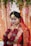 A person dressed in traditional wedding attire adorned with intricate gold jewelry and a red and gold saree. The headpiece is ornate and white, adding to the elegance. The backdrop features floral decorations and soft lighting, enhancing the ceremonial atmosphere.