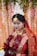 A person dressed in traditional wedding attire adorned with intricate gold jewelry and a red and gold saree. The headpiece is ornate and white, adding to the elegance. The backdrop features floral decorations and soft lighting, enhancing the ceremonial atmosphere.