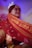 A person wearing traditional attire is visible, with a focus on an intricately embroidered red and gold veil. The veil features detailed patterns and script. The background indicates an indoor setting with some blurred decorative elements.