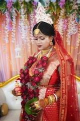 A bride is dressed in traditional red attire, adorned with intricate gold jewelry and a floral headpiece. She is holding a garland of red roses and green leaves, with her hands decorated with henna. Behind her, there is a backdrop of hanging flowers and leaves, creating a vibrant and elegant setting.