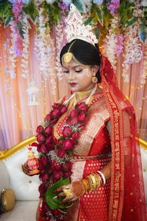 A bride is dressed in traditional red attire, adorned with intricate gold jewelry and a floral headpiece. She is holding a garland of red roses and green leaves, with her hands decorated with henna. Behind her, there is a backdrop of hanging flowers and leaves, creating a vibrant and elegant setting.