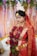 A bride is dressed in traditional red attire, adorned with intricate gold jewelry and a floral headpiece. She is holding a garland of red roses and green leaves, with her hands decorated with henna. Behind her, there is a backdrop of hanging flowers and leaves, creating a vibrant and elegant setting.