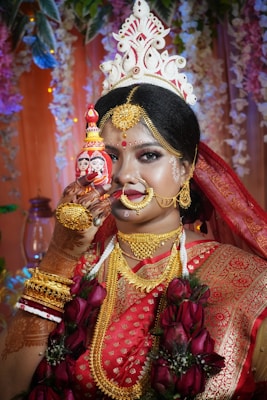 A woman dressed in traditional attire, adorned with elaborate jewelry and intricate henna designs on her hands. She wears a richly embroidered red and gold sari and holds a small decorative object with painted faces. Her elaborate headdress and vibrant floral garland add to the festive and ceremonial atmosphere.