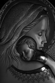 A detailed metal or embossed artwork depicting a woman holding a sleeping child. The intricate designs suggest a halo around the woman's head, and there are ornate patterns in her garment and headdress. The image conveys tenderness and serenity.