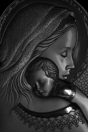A detailed metal or embossed artwork depicting a woman holding a sleeping child. The intricate designs suggest a halo around the woman's head, and there are ornate patterns in her garment and headdress. The image conveys tenderness and serenity.
