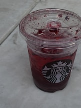 A cold beverage in a clear plastic cup with a logo, filled with a dark red liquid and ice, resting on a white marble surface. The lid is splattered with reddish fruit pieces or syrup.
