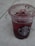 A cold beverage in a clear plastic cup with a logo, filled with a dark red liquid and ice, resting on a white marble surface. The lid is splattered with reddish fruit pieces or syrup.