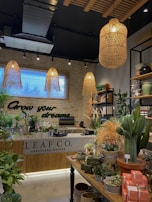 A lush indoor plant shop with a variety of greenery displayed on tables and shelves. The shop features an earthy and welcoming design, with wooden elements and wicker light fixtures hanging from the ceiling. A neon sign on the brick wall reads 'Grow your dreams...' adding a motivational touch. The space is filled with different types of plants, pots, and garden accessories, creating a vibrant and inviting atmosphere.
