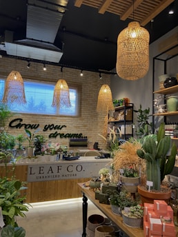 A lush indoor plant shop with a variety of greenery displayed on tables and shelves. The shop features an earthy and welcoming design, with wooden elements and wicker light fixtures hanging from the ceiling. A neon sign on the brick wall reads 'Grow your dreams...' adding a motivational touch. The space is filled with different types of plants, pots, and garden accessories, creating a vibrant and inviting atmosphere.