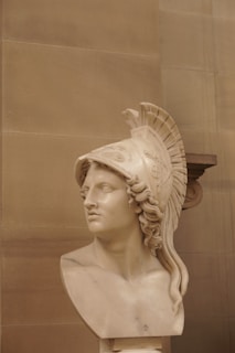 A marble bust sculpture depicting a classical warrior wearing a detailed helmet with a crest. The expression on the face appears focused and noble. The background is composed of simple, light-colored stone tiles.