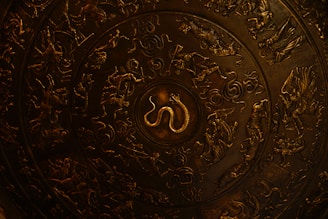 A decorative circular metal plate featuring intricate engravings of mythical creatures and figures, including a prominent snake at the center surrounded by people, animals, and swirling patterns.
