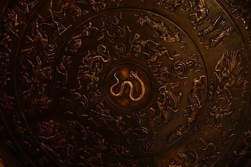 A decorative circular metal plate featuring intricate engravings of mythical creatures and figures, including a prominent snake at the center surrounded by people, animals, and swirling patterns.