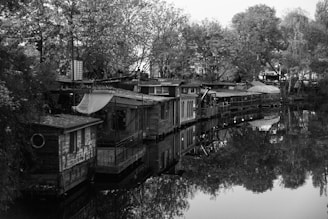 A row of rustic houseboats lined up along the edge of a calm body of water, surrounded by lush trees. The scene is captured in grayscale, emphasizing the textures of the wood and the reflections on the water's surface. The structures appear to be handmade, with various makeshift additions like tarps and panels, suggesting a bohemian lifestyle.