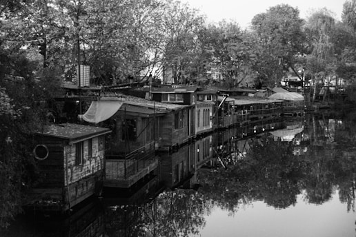 A row of rustic houseboats lined up along the edge of a calm body of water, surrounded by lush trees. The scene is captured in grayscale, emphasizing the textures of the wood and the reflections on the water's surface. The structures appear to be handmade, with various makeshift additions like tarps and panels, suggesting a bohemian lifestyle.
