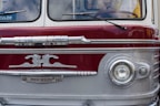 A vintage bus with a distinctive front design, featuring an emblem and retro headlights. The bus has a two-toned color scheme with maroon and silver hues and chrome accents. Visible portions of the interior show the steering wheel and a partial view of a person inside.